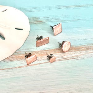 May include: Five pairs of copper-colored stud earrings in various geometric shapes: square, rectangle, circle, and oval. The earrings have a hammered texture and are displayed on a light blue and white wooden surface.