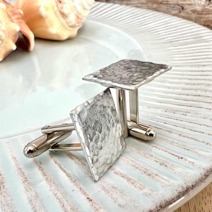 May include: A pair of silver cufflinks with a hammered texture, displayed on a light blue, ridged plate. The cufflinks have a square shape and a shiny finish. Seashells are visible in the background.