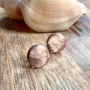 May include: Two copper-colored, round stud earrings with a textured surface. The earrings are displayed on a wooden surface, with a seashell in the background. The earrings have a brushed metal appearance.