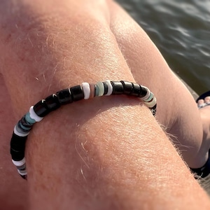 May include: A black and white beaded bracelet with a few green and blue beads. The bracelet is worn on a person's wrist.