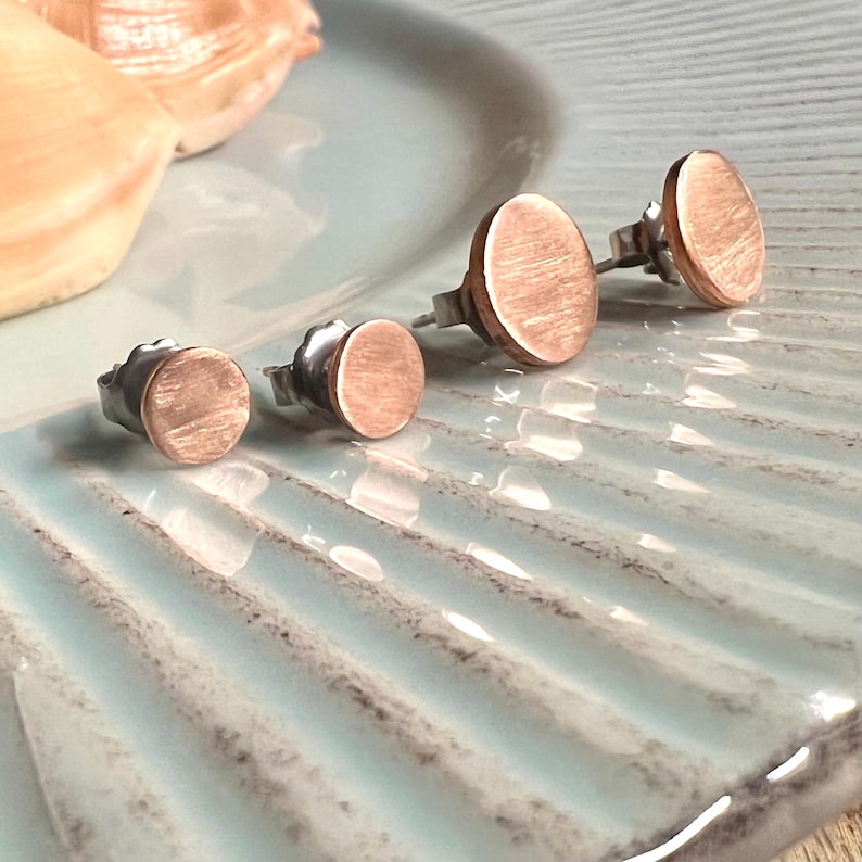 May include: Four pairs of copper-colored stud earrings with a brushed metal finish. The earrings are circular and vary in size, displayed on a light blue, textured surface. The earrings have silver posts and backs.