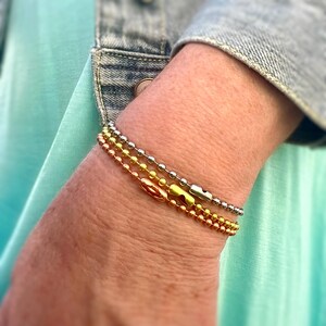 May include: A set of three bracelets with different colored beads. The bracelets are made of metal and have a silver, gold, and rose gold finish. The bracelets are stacked on a person's wrist.