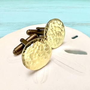 May include: A pair of gold-toned cufflinks with a hammered texture. The cufflinks are round and have a shiny finish.