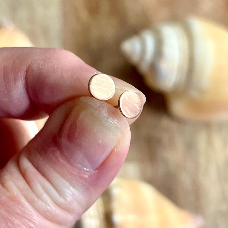 May include: A pair of small, round stud earrings. The earrings are a light, warm color, possibly rose gold or copper. The studs are held between fingers, with a seashell in the background.