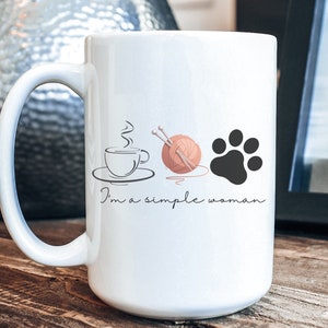 May include: White ceramic mug with a large handle, featuring illustrations of a coffee cup, yarn with knitting needles, and a paw print. The text "I'm a simple woman" is written below the illustrations. The mug is on a wooden surface.