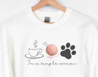 May include: White sweatshirt with a graphic design of a coffee cup, yarn and knitting needles, and a paw print. The text "I'm a simple woman" is printed below the graphic.