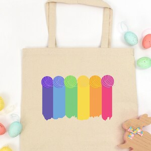 May include: A beige tote bag with a rainbow yarn ball design. The bag has two handles and is made of canvas. The bag is surrounded by pastel colored Easter eggs.