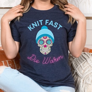 May include: A navy blue t-shirt with the text "Knit Fast Die Warm" and a sugar skull wearing a blue knit hat.