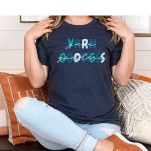 May include: A woman wearing a navy blue t-shirt with the text "yarn goddess" in colorful lettering. She is sitting on a bench with a white and orange pillow behind her.