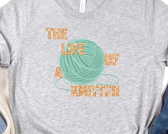 May include: A light gray t-shirt with the text "THE LIFE OF A KNITTER" in gold glitter lettering. A teal yarn ball graphic is in the center. The shirt is paired with blue jeans and a cream-colored sweater.