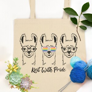 May include: A beige tote bag with a black and white illustration of three llamas wearing glasses. The middle llama is wearing rainbow sunglasses. The bag has the text "Knit With Pride" printed on it. The bag is surrounded by yarn balls and knitting needles.