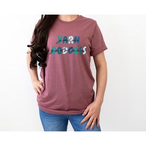 May include: A mauve t-shirt with the text "Yarn Goddess" in colorful, stylized lettering.