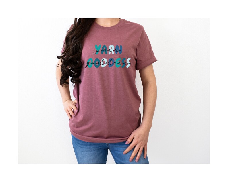 May include: A mauve t-shirt with the text "Yarn Goddess" in colorful, stylized lettering.