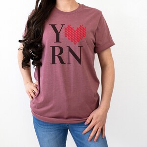 May include: A mauve t-shirt with the text "Y" followed by a red knitted heart and the text "RN".