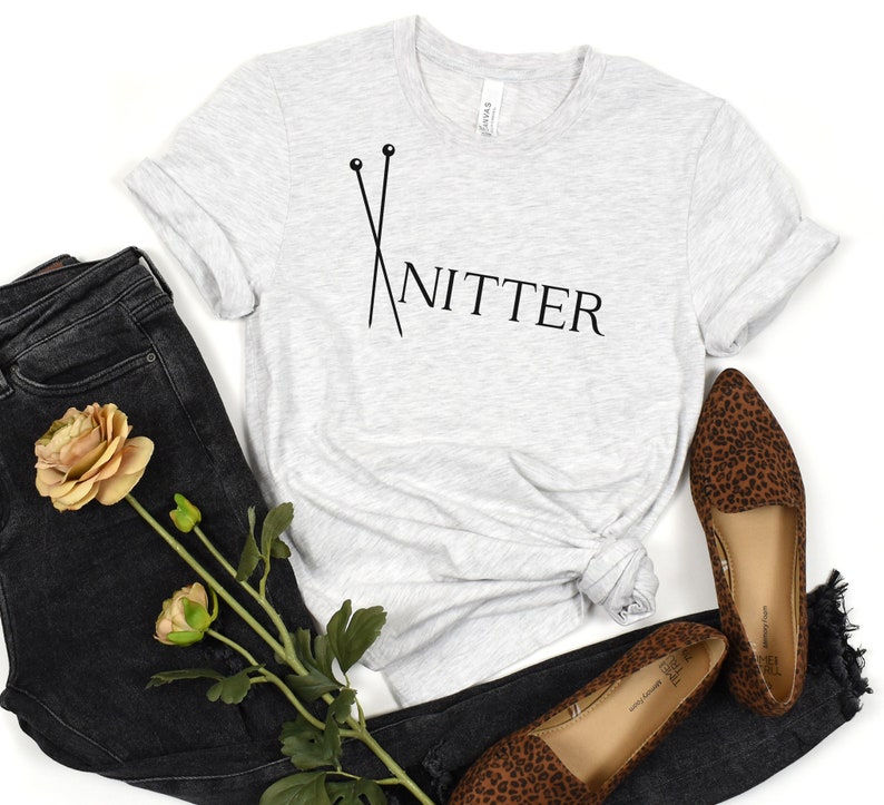 May include: A gray t-shirt with a black graphic of knitting needles and the word "Knitter" on it. The shirt is laid out on a white background with a pair of brown and black leopard print shoes and a pair of black jeans with a brown flower.
