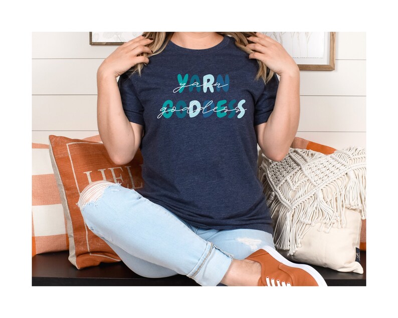 May include: A woman wearing a navy blue t-shirt with the text "yarn goddess" in colorful lettering. She is sitting on a bench with a white and orange pillow behind her.