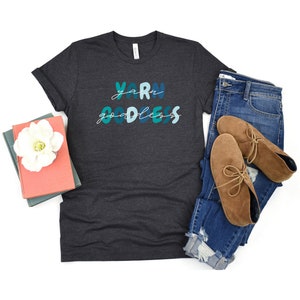 May include: A dark gray t-shirt with the text "Yarn Goddess" in blue and green lettering. The shirt is paired with blue jeans and brown ankle boots.
