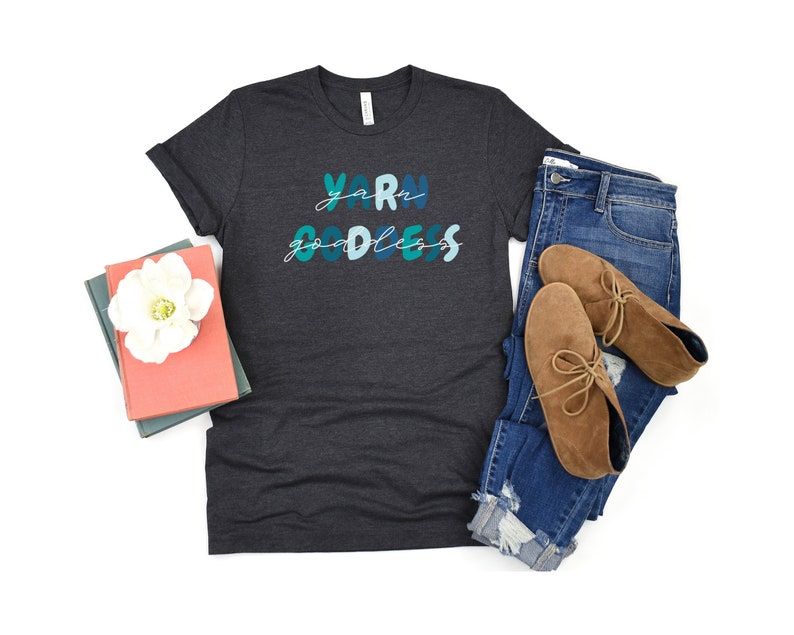May include: A dark gray t-shirt with the text "Yarn Goddess" in blue and green lettering. The shirt is paired with blue jeans and brown ankle boots.