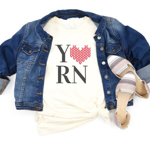 May include: A white t-shirt with the text "Y" followed by a red knitted heart and the text "RN". The shirt is worn over a denim jacket and paired with striped sandals.