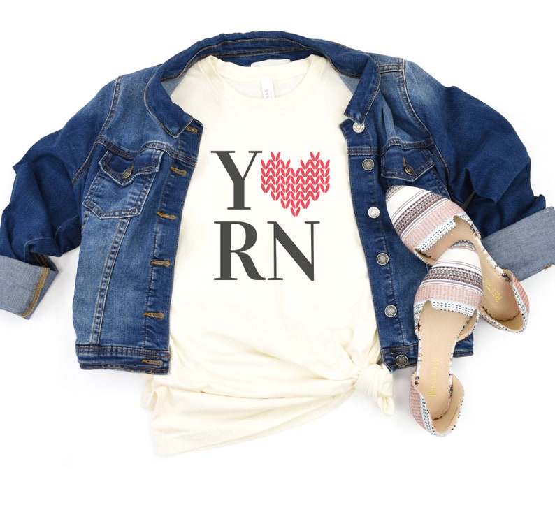 May include: A white t-shirt with the text "Y" followed by a red knitted heart and the text "RN". The shirt is worn over a denim jacket and paired with striped sandals.