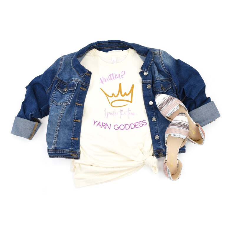 May include: A cream colored t-shirt with the text "Knitter? I prefer the term... Yarn Goddess" in purple and gold. A gold crown graphic is above the text. The shirt is worn with a blue denim jacket and striped sandals.