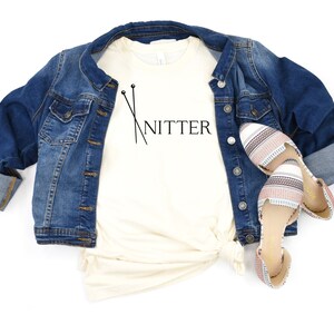 May include: A white t-shirt with the text "Knitter" and a pair of knitting needles printed on it. The shirt is paired with a denim jacket and striped flats.