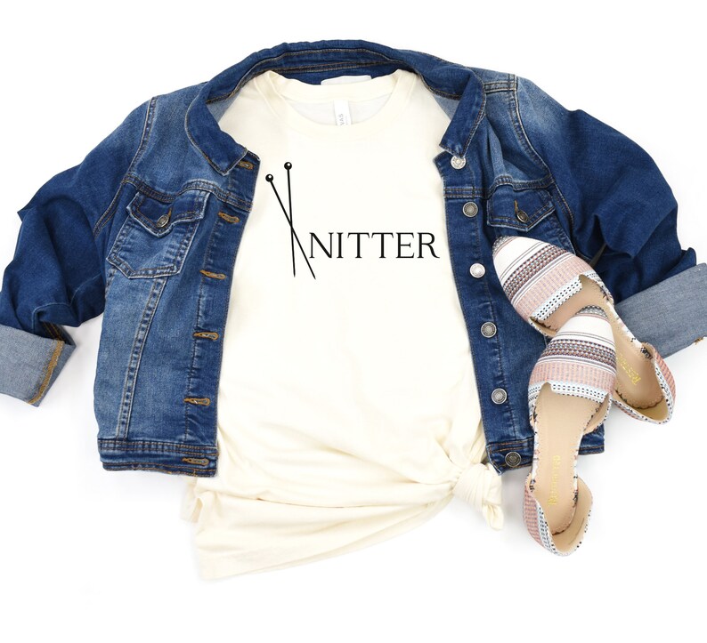 May include: A white t-shirt with the text "Knitter" and a pair of knitting needles printed on it. The shirt is paired with a denim jacket and striped flats.