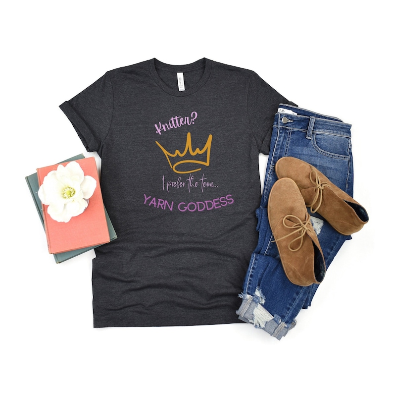 May include: A dark gray t-shirt with the text "Knitter? I prefer the term... YARN GODDESS" in pink and gold lettering. A gold crown graphic is above the text.