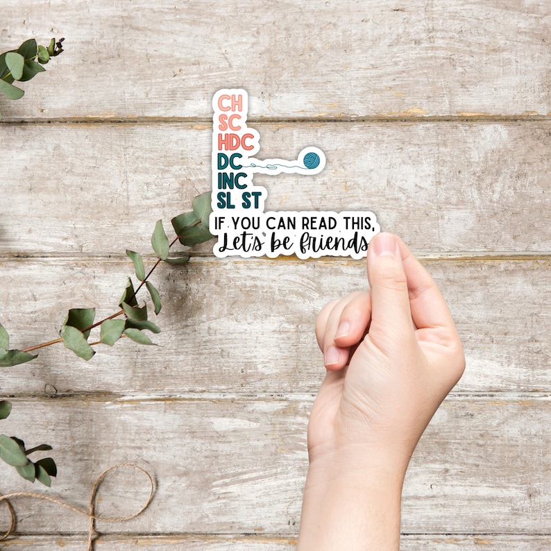 May include: A white sticker with pink and teal text listing crochet abbreviations and the phrase "If you can read this, Let's be friends." A teal yarn ball graphic is included. The sticker is held by a hand.
