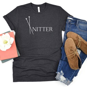 May include: A dark gray t-shirt with a white graphic of knitting needles and the word "Knitter". The shirt is shown on a white background with a pair of blue jeans and brown ankle boots.