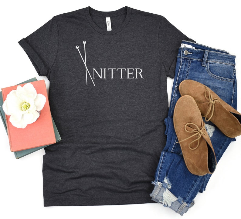 May include: A dark gray t-shirt with a white graphic of knitting needles and the word "Knitter". The shirt is shown on a white background with a pair of blue jeans and brown ankle boots.