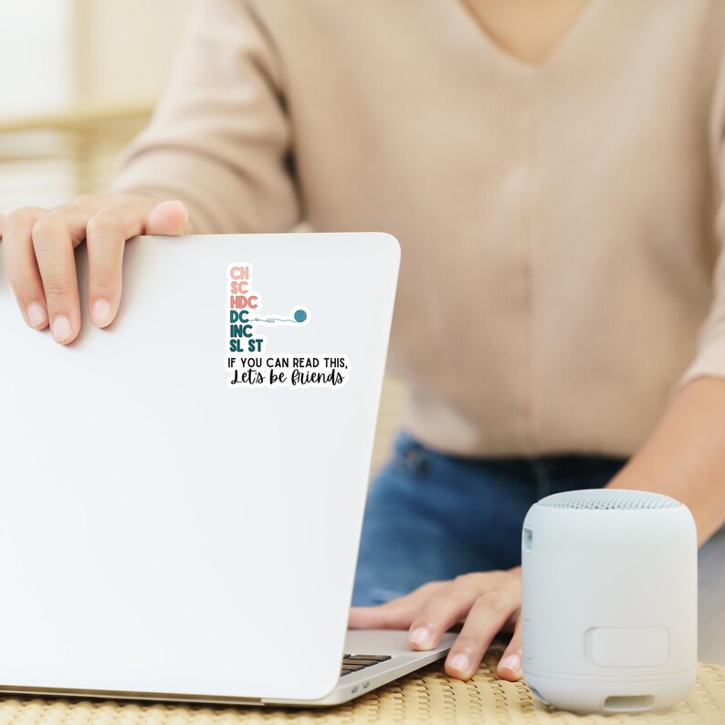 May include: A white laptop with a sticker that reads "CH, SC, HDC, DC, INC, SL ST. If you can read this, Let's be friends." A small, white, cylindrical speaker sits to the right of the laptop.
