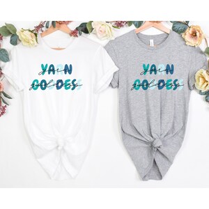 May include: Two t-shirts with the text "Yarn Goddess" in colorful letters. The shirts are white and grey and are hanging on hangers.