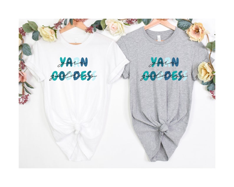 May include: Two t-shirts with the text "Yarn Goddess" in colorful letters. The shirts are white and grey and are hanging on hangers.