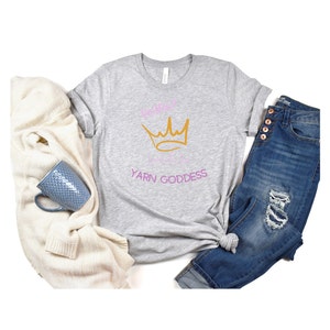 May include: A heather grey t-shirt with a pink and gold crown and the text "Remarkable Yarn Goddess".