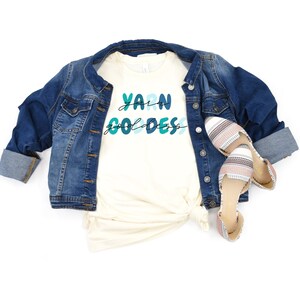 May include: A white t-shirt with the text "Yarn Goddess" in blue and green lettering. The shirt is paired with a denim jacket and striped flats.