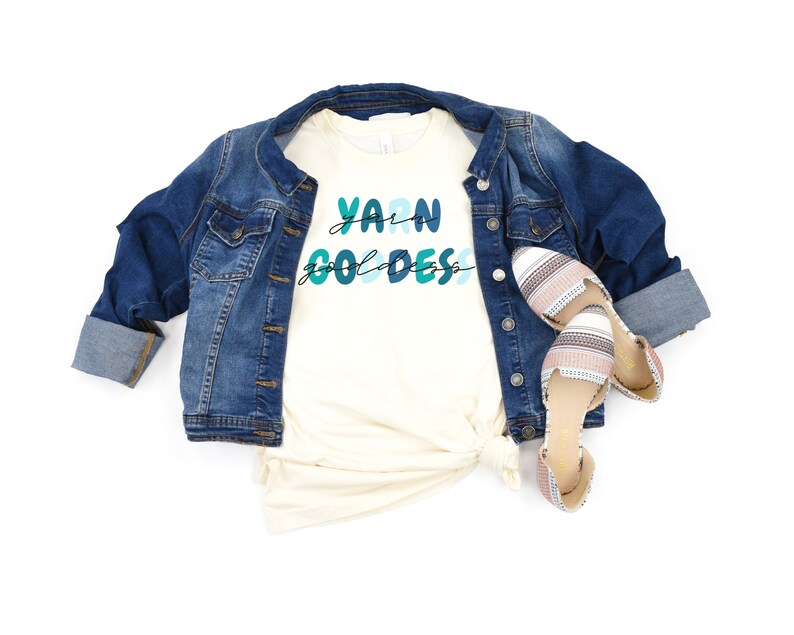 May include: A white t-shirt with the text "Yarn Goddess" in blue and green lettering. The shirt is paired with a denim jacket and striped flats.