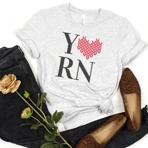 May include: A gray t-shirt with the text "YRN" in black letters and a red knitted heart in the center. The heart is made of yarn and has a white outline.
