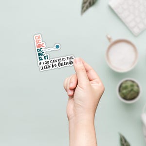 May include: A white sticker with teal and pink text that reads "CH, SC, HDC, DC, INC, SL ST, IF YOU CAN READ THIS, Let's be friends." The sticker is held by a hand against a light green background.