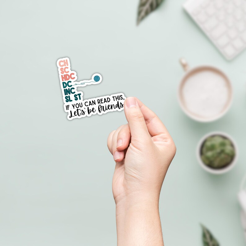 May include: A white sticker with teal and pink text that reads "CH, SC, HDC, DC, INC, SL ST, IF YOU CAN READ THIS, Let's be friends." The sticker is held by a hand against a light green background.