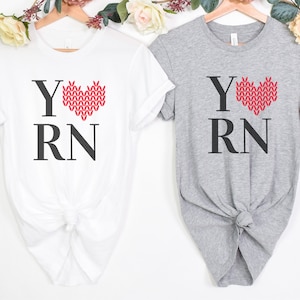 May include: Two t-shirts with the text "Y RN" and a red knitted heart. The heart is a stylized representation of a knitted stitch. One shirt is white and the other is gray.
