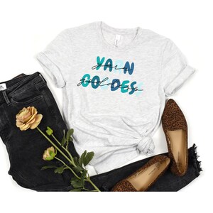 May include: A gray t-shirt with the text "Yarn Goddess" in blue and green lettering. The shirt is paired with black jeans and leopard print flats.