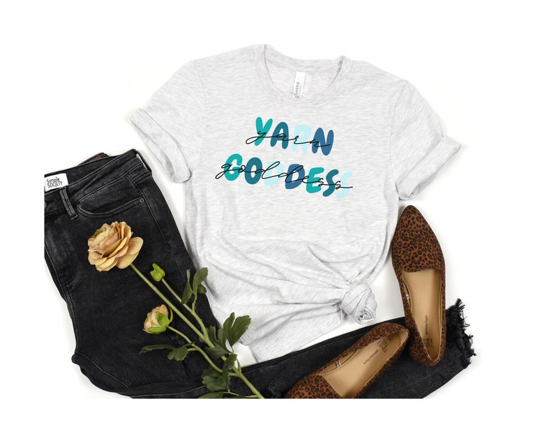 May include: A gray t-shirt with the text "Yarn Goddess" in blue and green lettering. The shirt is paired with black jeans and leopard print flats.