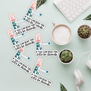 May include: A collection of white stickers with the text "IF YOU CAN READ THIS, Let's be friends" and crochet abbreviations. The stickers are scattered on a light green surface, alongside a cup of coffee, succulents, and a keyboard.