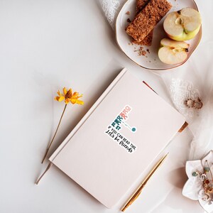 May include: A white journal with a sticker that reads "If you can read this, let's be friends." A gold pen, a yellow flower, and a plate with apple slices and a granola bar are also visible.