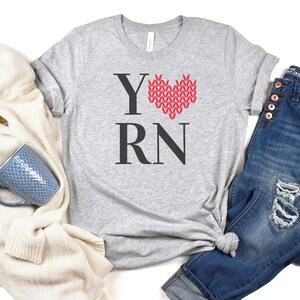 May include: A gray t-shirt with the text "Y RN" and a red knitted heart design. The heart is in the shape of a love heart.