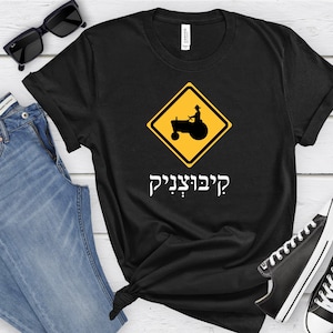 Może przedstawiać: Czarny T-shirt z żółtym znakiem ostrzegawczym przedstawiającym traktor i tekst "?דברגוניק" w języku hebrajskim.