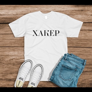 May include: White t-shirt with the word "XAKEP" printed in black on the front. The shirt is laid flat on a wooden surface with a pair of white sneakers and blue jeans.
