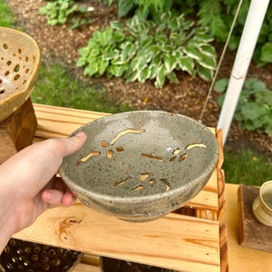 May include: A handmade ceramic bowl with a speckled gray glaze and decorative cut-out patterns. The bowl is held in a person's hand and sits on a wooden shelf with other pottery. The bowl's diameter is approximately 8 inches.