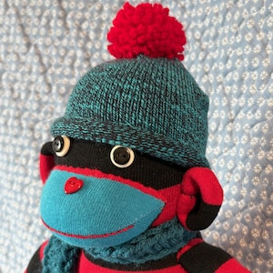 May include: A handmade sock monkey doll wearing a teal and black knit hat with a red pom-pom. The monkey has a teal face, red ears, and a blue scarf. The body is red and black striped.